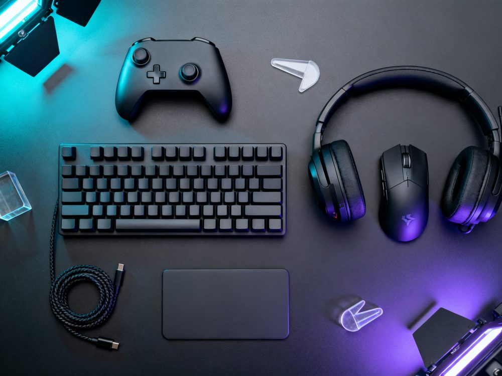 Equipamento gaming com teclado, comando preto, rato e auscultadores dispostos sobre superfície escura com luz LED azul e roxa