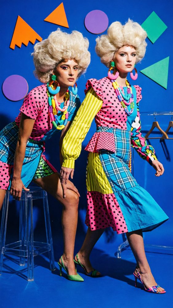 Duas modelos com roupas patchwork coloridas e acessórios grandes em fundo azul com formas geométricas