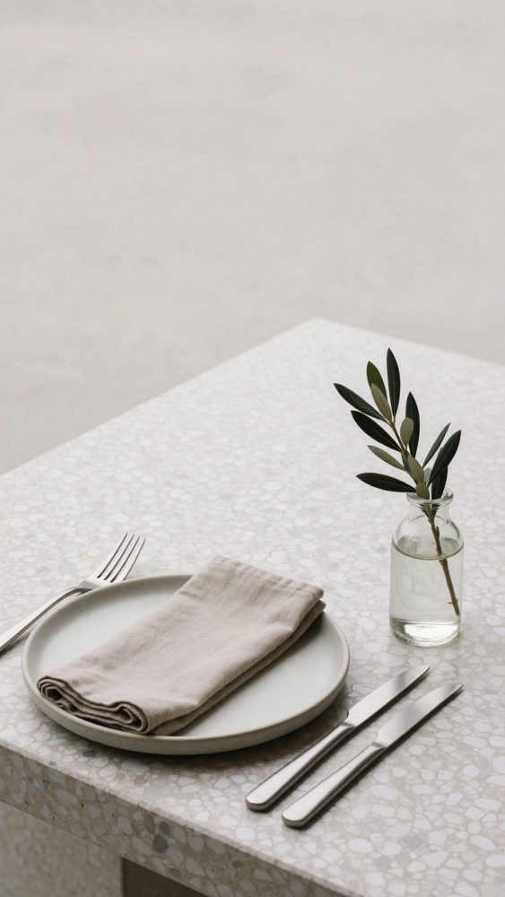 Mesa minimalista com prato branco, guardanapo bege, talheres prateados e ramo verde em jarrinha de vidro
