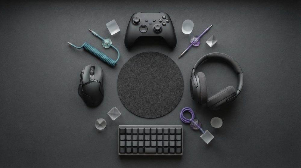 Acessórios de gaming negro com teclado, rato, comando e auscultadores dispostos sobre fundo escuro