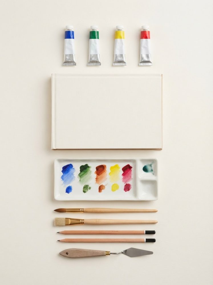 Materiais de pintura organizados com tubos de tinta, pincéis, paleta, lápis, espátula e tela em branco