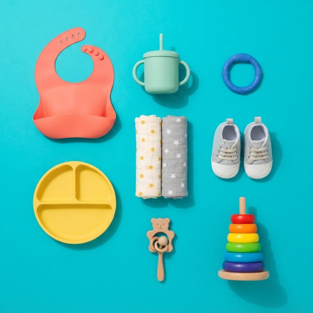 Itens variados para bebé dispostos sobre fundo azul, incluindo babete, copo e brinquedos coloridos