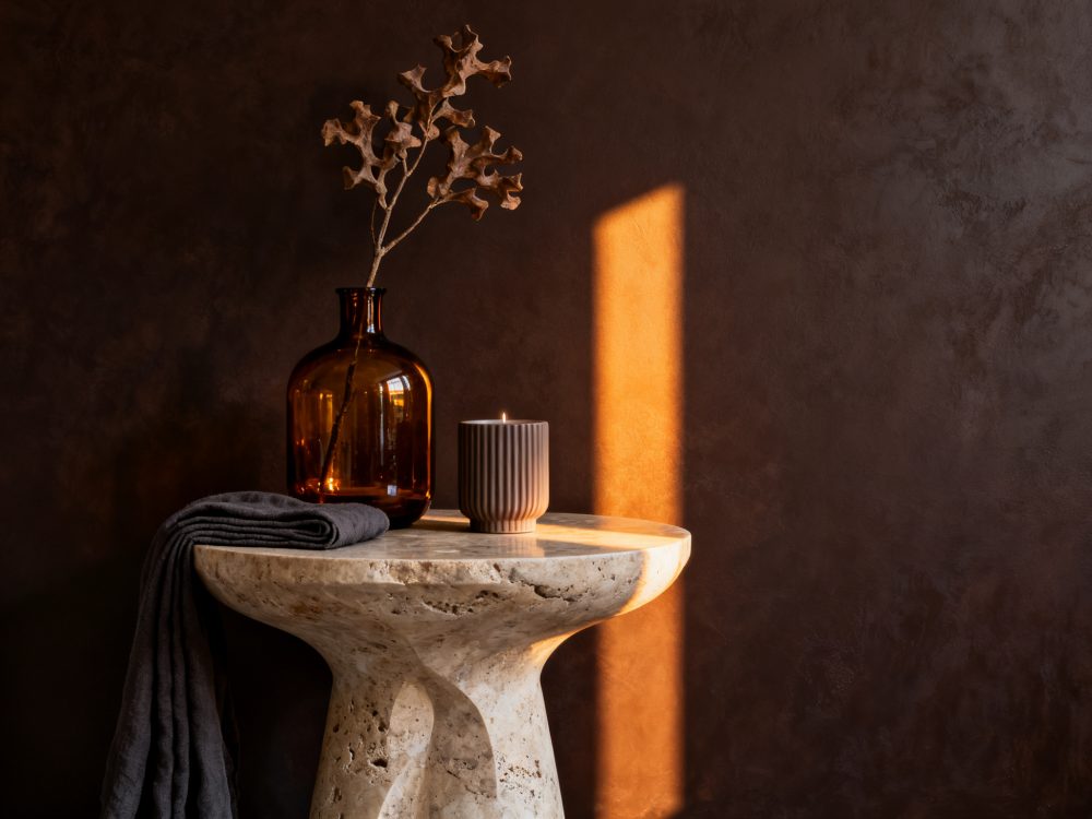 Mesa de pedra com jarra âmbar, flores secas, vela e tecido cinza com luz natural quente