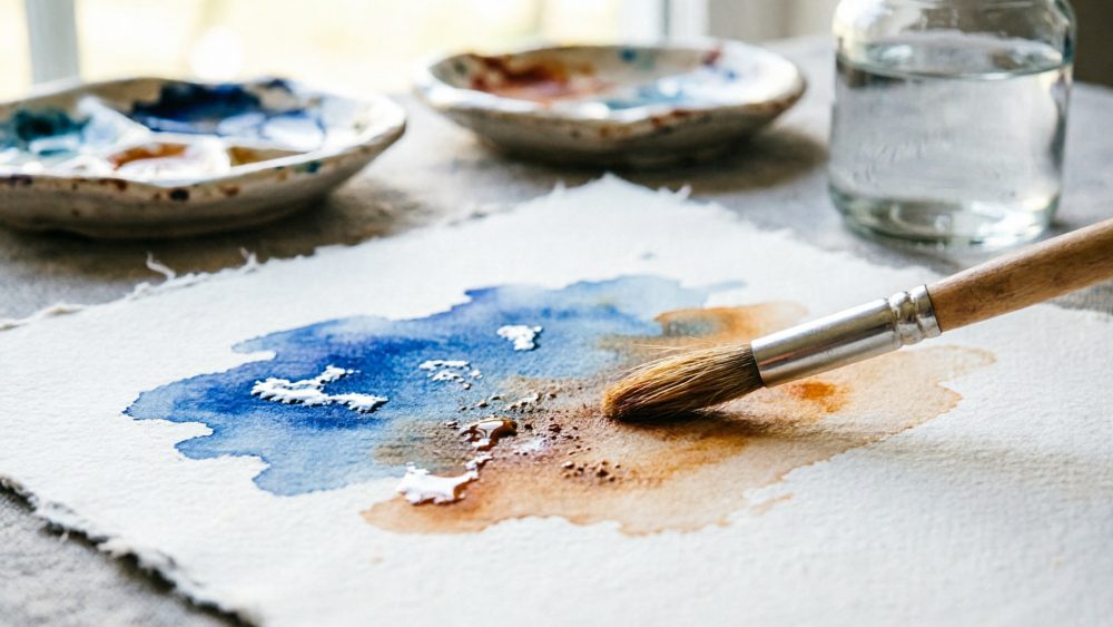 Pincel molhado a aplicar tintas azuis e castanhas em papel para aquarela junto a paletas e frasco de água