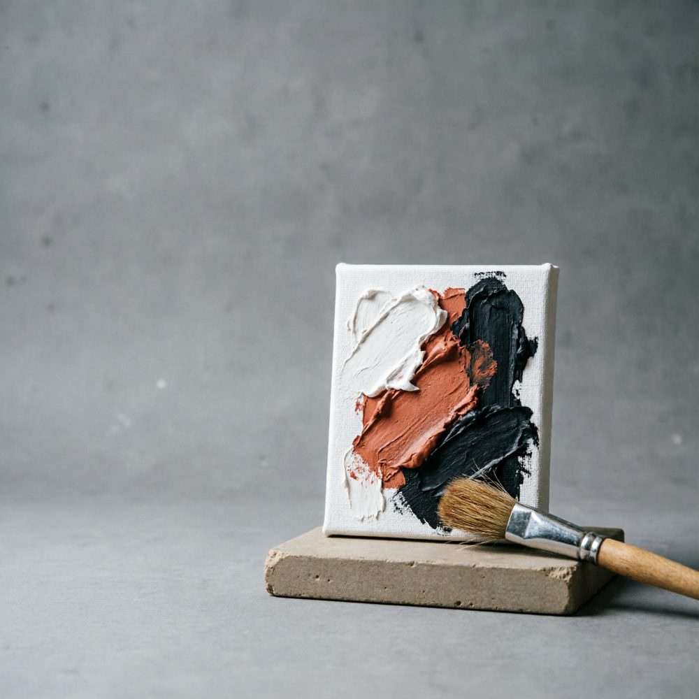 Mini tela com pintura a óleo em branco, terracota e preto, pincel sobre base cinzenta