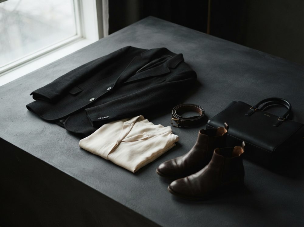 Conjunto de roupa com blazer preto, camisa clara, cinto castanho, botas de pele e mala preta sobre superfície escura