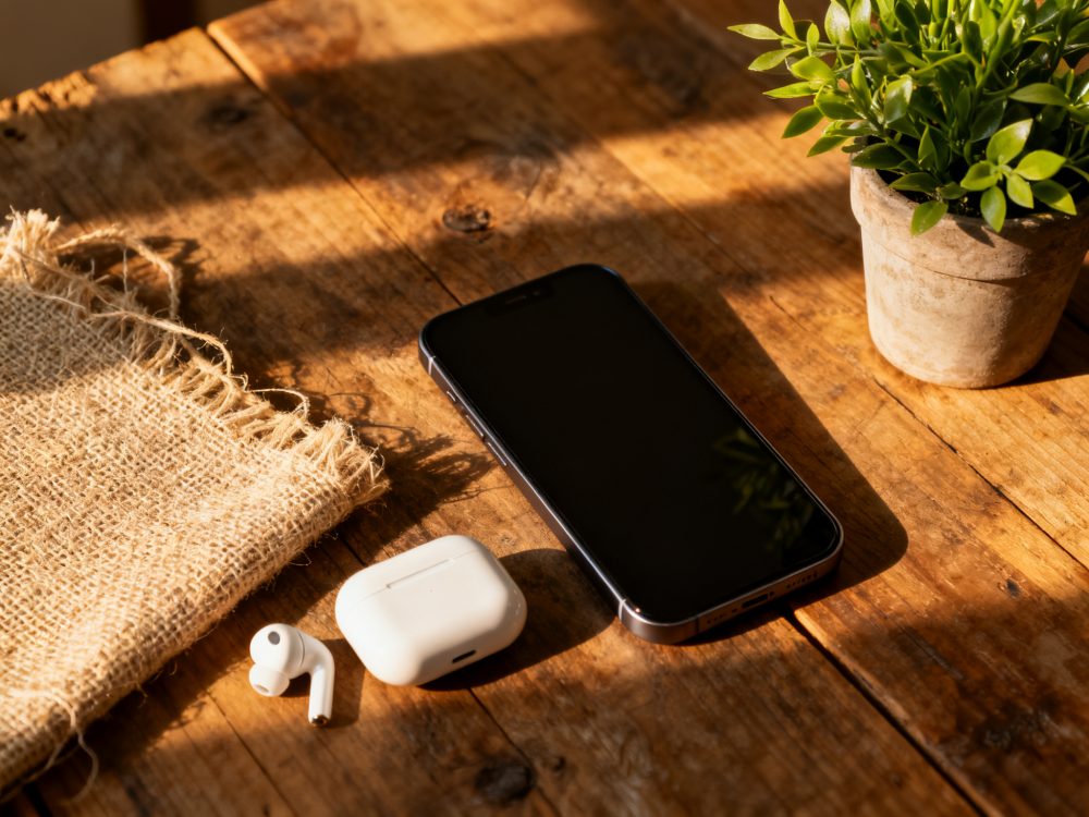 Smartphone e earbuds brancos numa mesa de madeira com luz natural, planta em vaso e tecido rústico