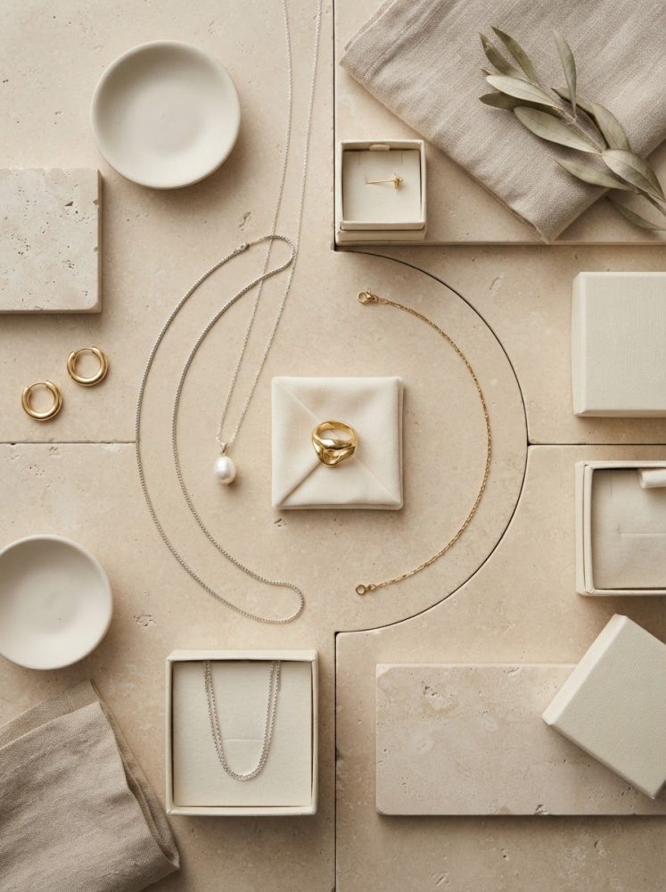 Joalharia minimalista com anéis dourados, colar com pérola e brincos em superfície neutra