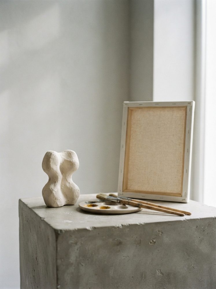 Pedestal de cimento com escultura abstrata, tela sem pintura e pincéis ao lado
