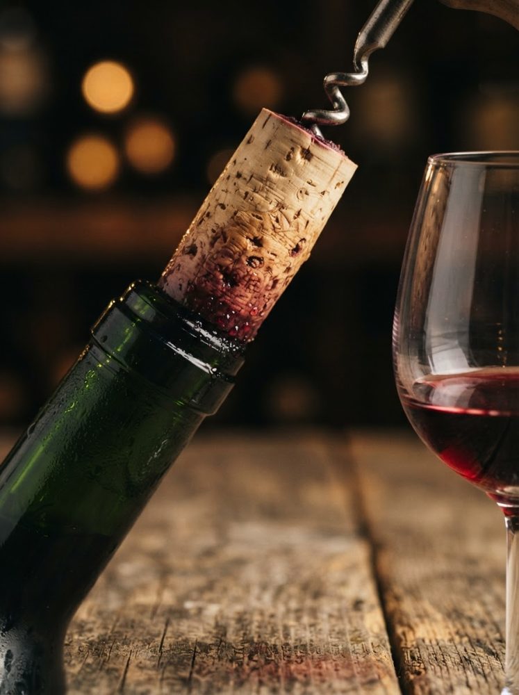Garrafa de vinho com rolha a ser retirada e copo com vinho tinto em superfície de madeira rústica