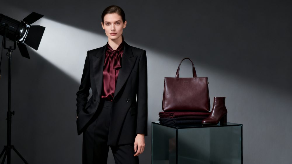 Mulher com blazer preto e camisa bordô, ao lado de mala e botas em couro bordô sobre suporte de vidro