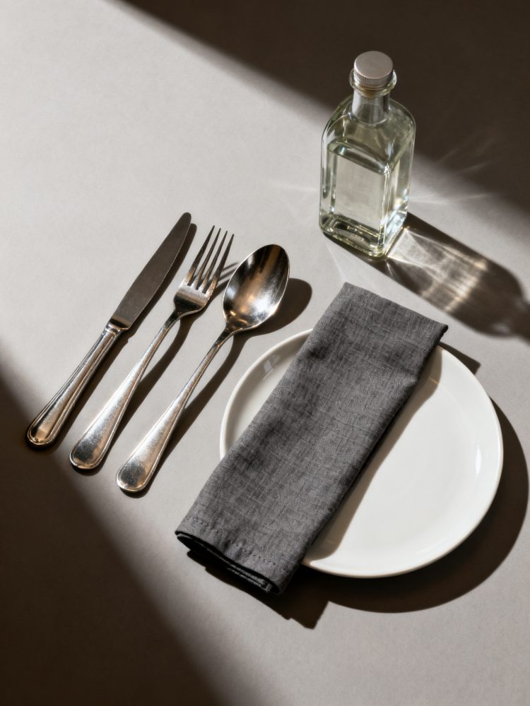 Talheres prateados com prato branco, guardanapo cinza escuro e garrafa de vidro sob luz natural