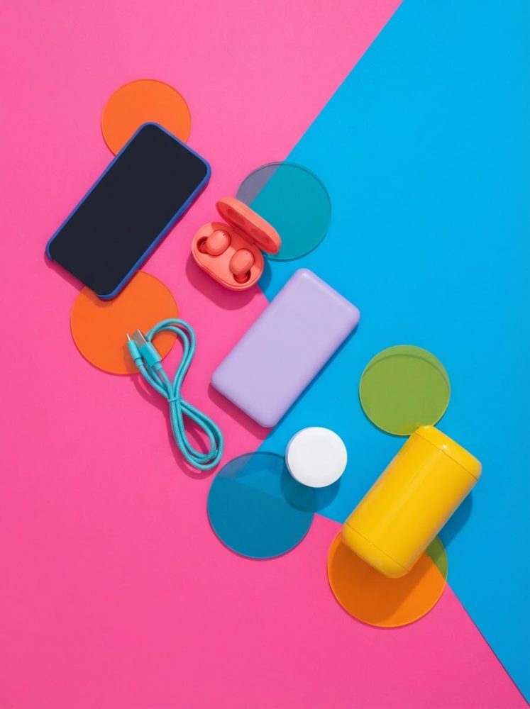 Smartphone, earbuds, power bank e cabo USB coloridos sobre fundo rosa e azul com círculos multicoloridos