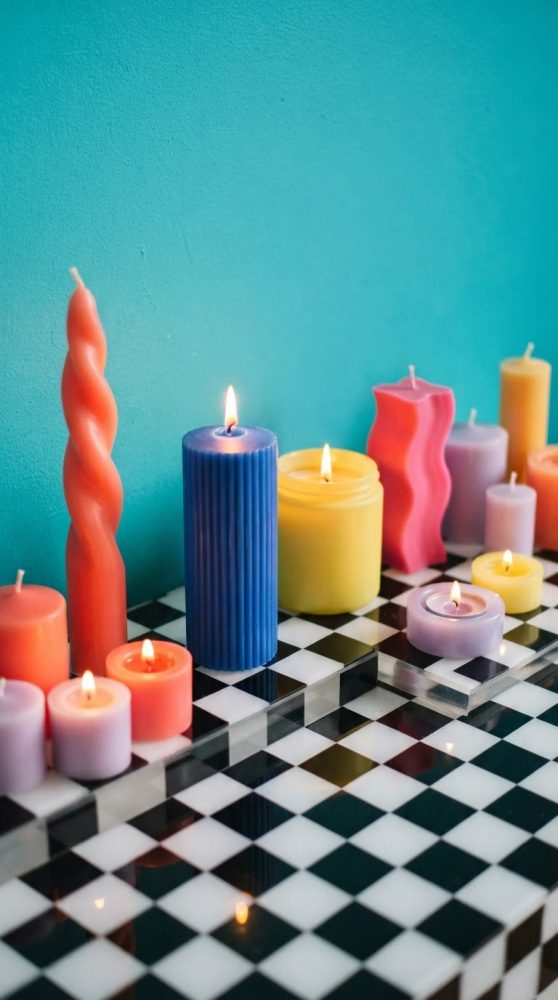 Várias velas coloridas em diferentes formas e algumas acesas sobre superfície quadriculada preta e branca
