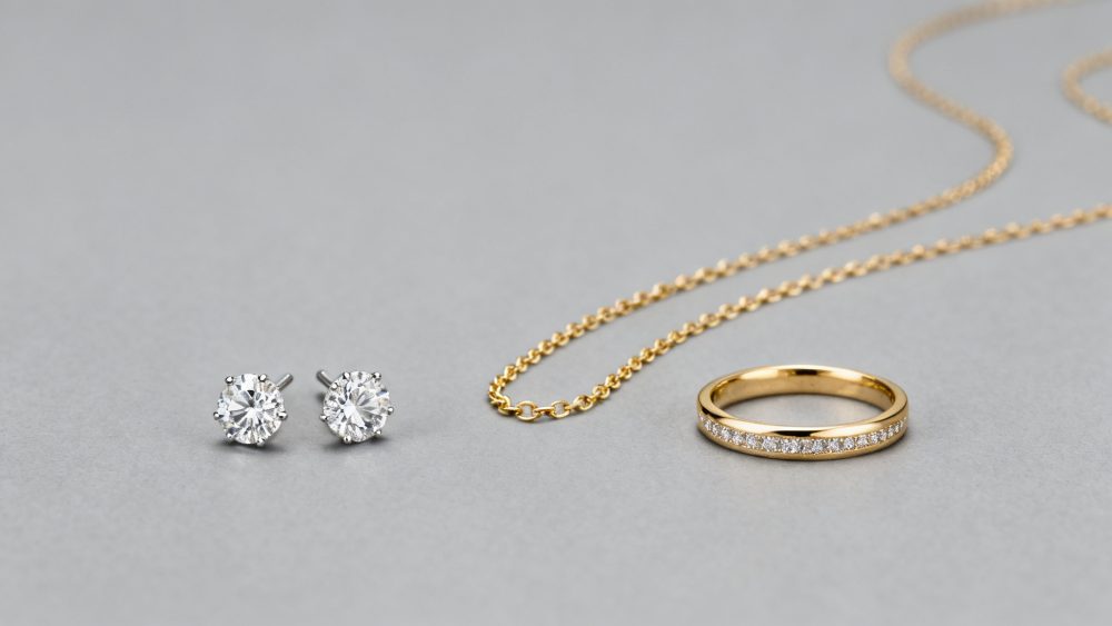 Anel dourado cravejado de diamantes, colar com corrente dourada e brincos de diamante sobre fundo cinzento