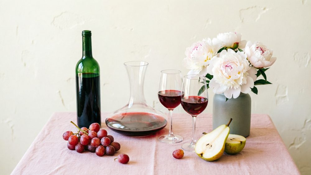 Mesa com garrafa e copos de vinho tinto, uvas, pêra cortada e vaso com flores brancas