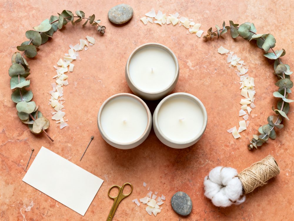 Três velas brancas em potes cerâmicos com decoração natural sobre superfície laranja texturada