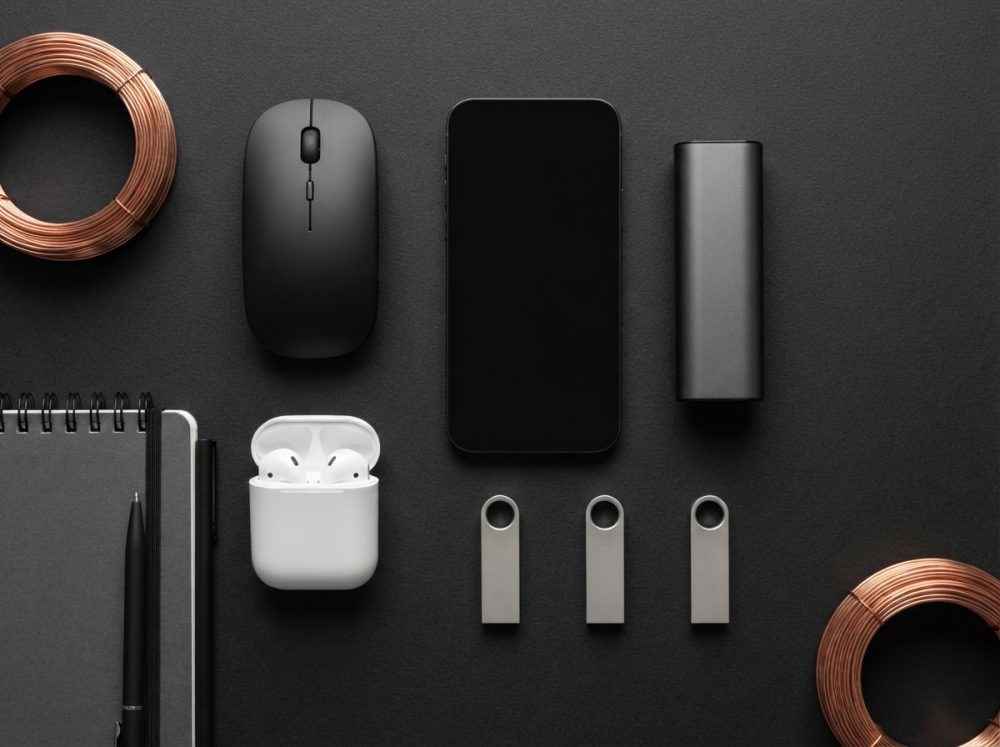 Acessórios tecnológicos organizados numa superfície escura com smartphone, rato, auriculares e pen drives