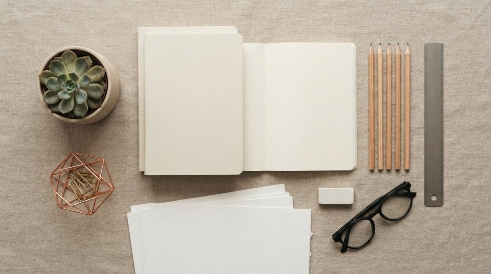 Material de papelaria minimalista com cadernos, lápis, régua, óculos, borracha e planta suculenta sobre fundo bege