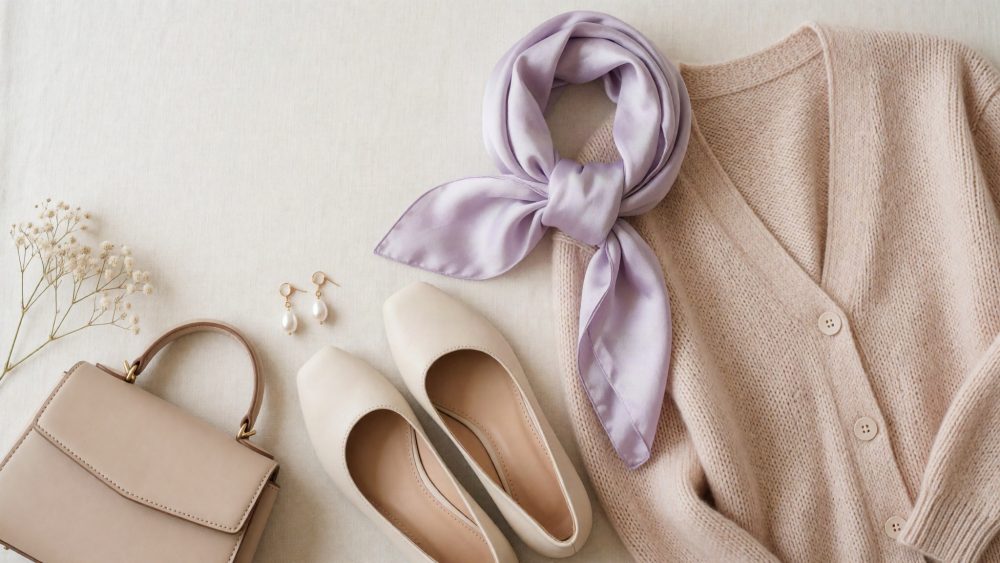 Cardigan bege, lenço lilás, sapatos de salto baixo, mala bege e brincos pérola dispostos numa superfície clara