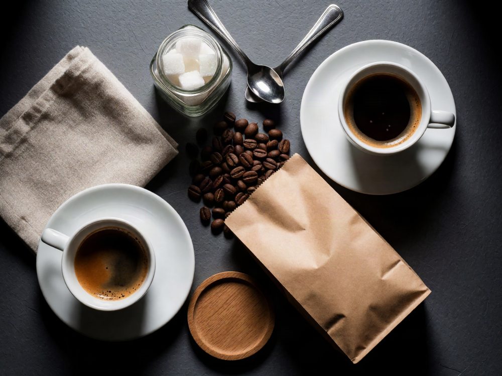 Duas chávenas de café, saco de papel com grãos, frasco de açúcar, colher e pano sobre mesa escura
