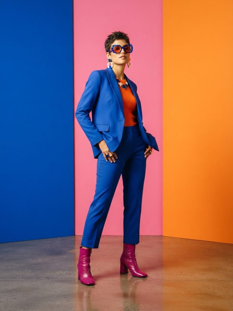 Mulher com fato azul, botas rosa e óculos grandes, em fundo azul, rosa e laranja