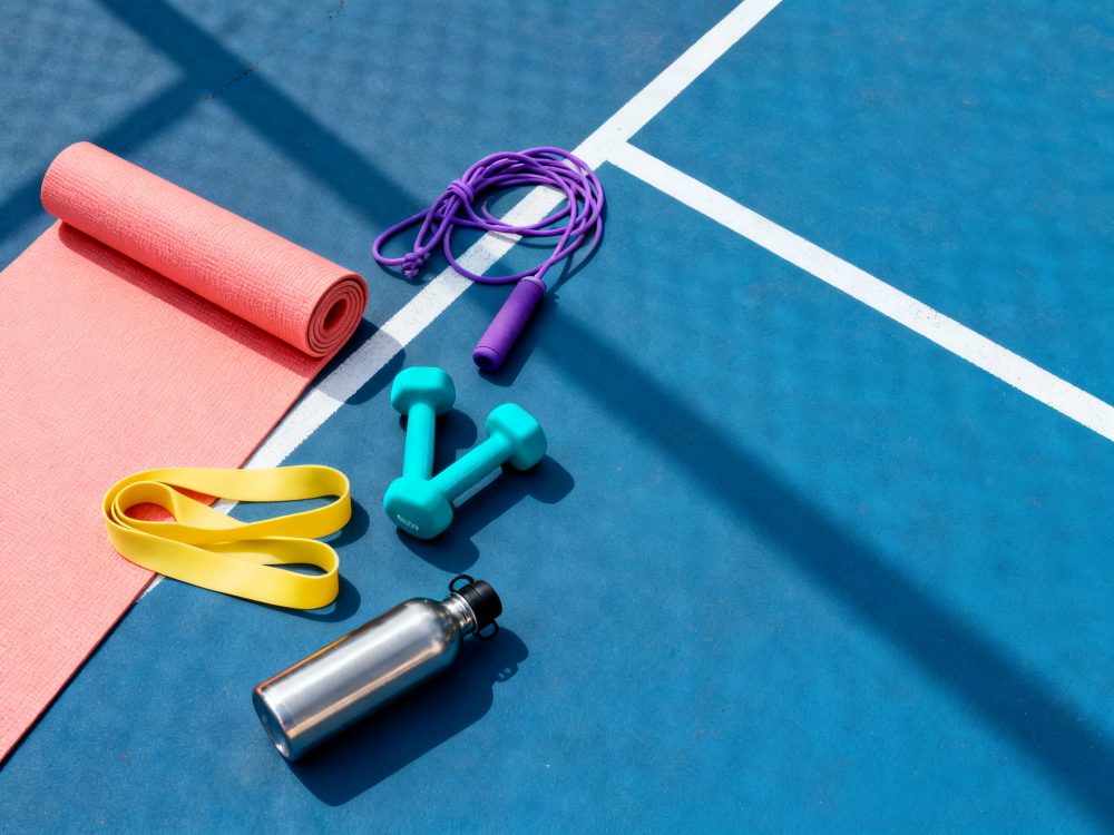Tapete de yoga, halteres, corda de pular, elástico fitness e garrafa em chão azul com linhas brancas