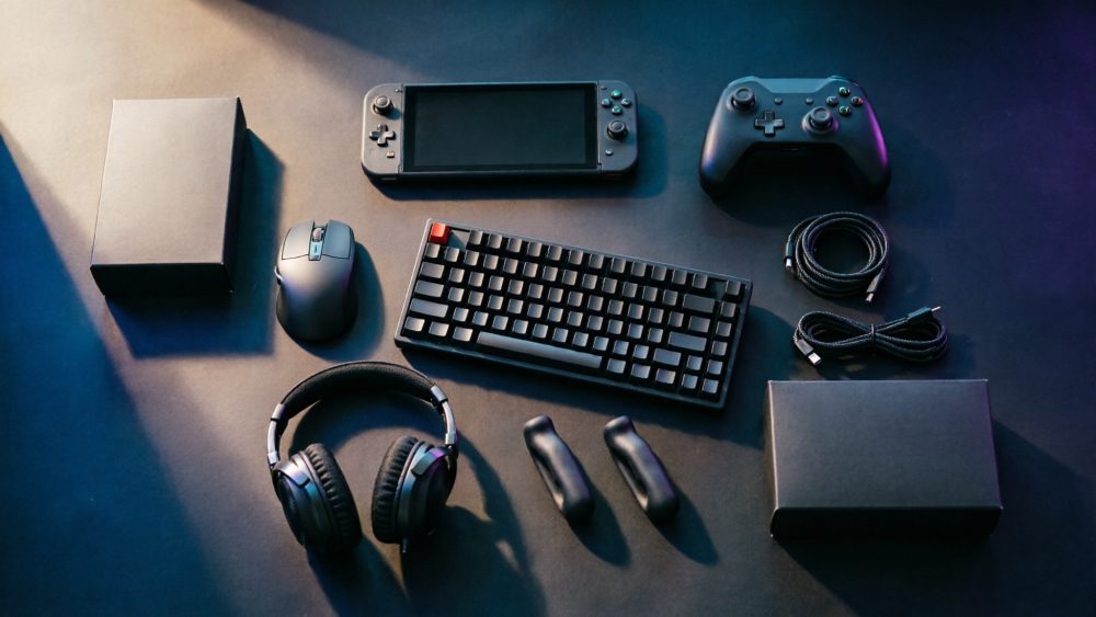 Acessórios gaming pretos, teclado, comandos, rato, auriculares e cabos sobre mesa escura