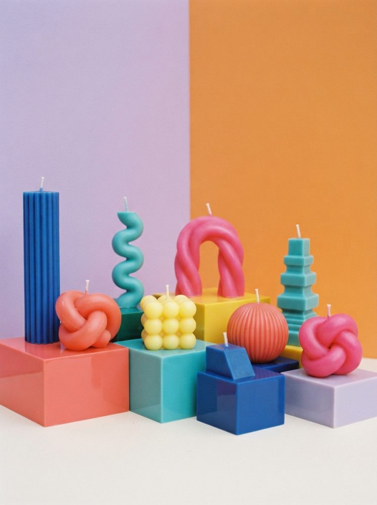 Velas coloridas em formas diversas sobre cubos de cores variadas com fundo lilás e laranja