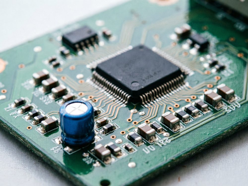Placa eletrónica verde com microchip e condensador azul em foco próximo
