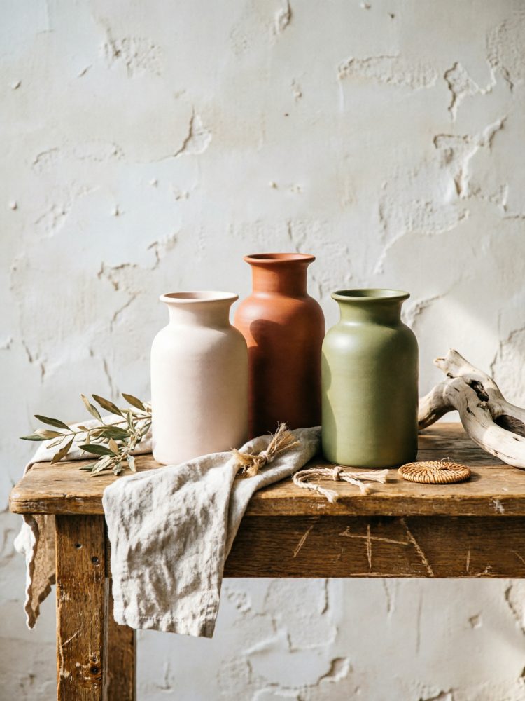 Três vasos cerâmicos mate bege, terracota e verde sobre mesa rústica com tecido e ramos secos