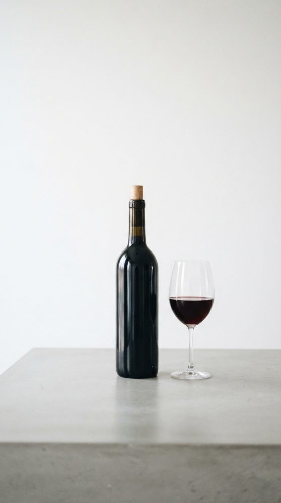 Garrafa de vinho tinto com rolha e copo de vinho parcial sobre mesa cinzenta