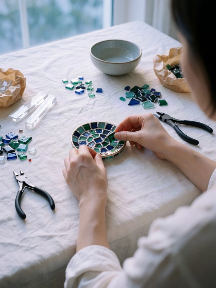 Mãos a colocar peças de vidro num mosaico colorido azul e verde sobre tecido branco com ferramentas perto