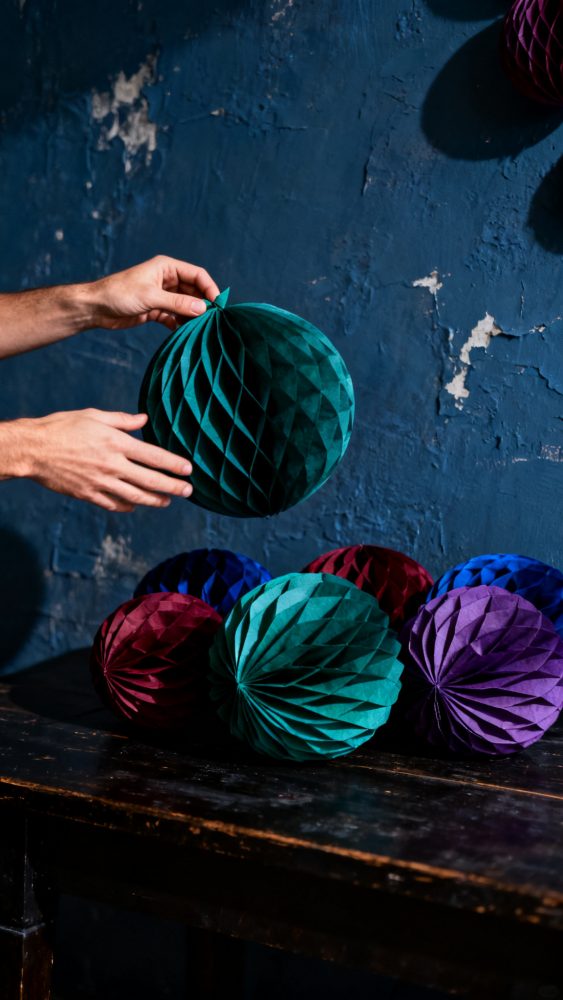 Mãos colocando bolas decorativas azuis, verdes, roxas e bordôs sobre uma mesa rústica com fundo azul gasto