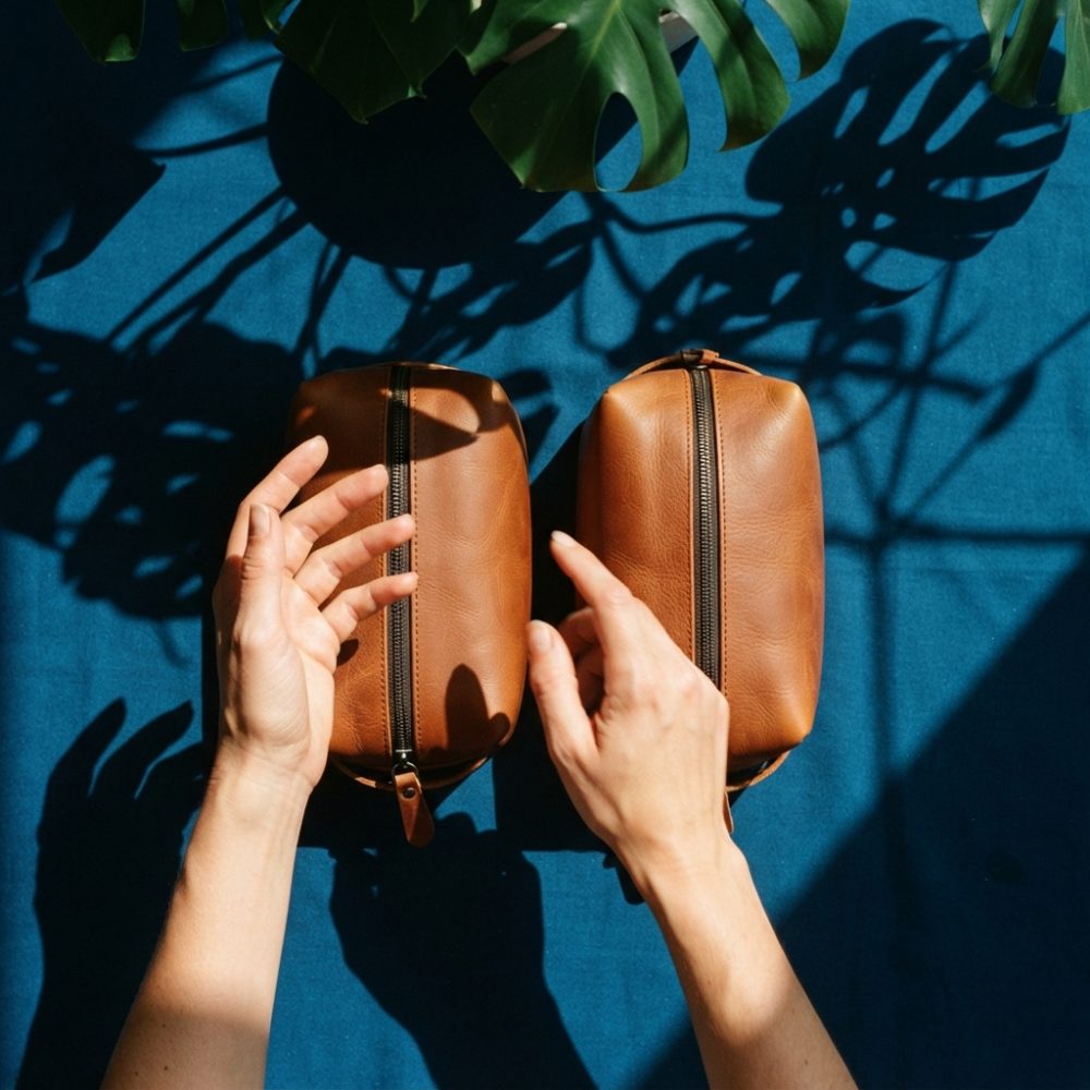 Duas necessaires castanhas sobre fundo azul com sombras de folhas tropicais e mãos femininas