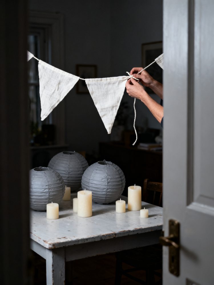 Mãos a prender bandeirolas de tecido branco com lanternas cinzentas e velas sobre mesa rústica