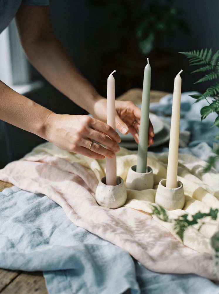 Mãos colocando velas coloridas em suportes de cerâmica sobre tecidos em tons pastel na mesa