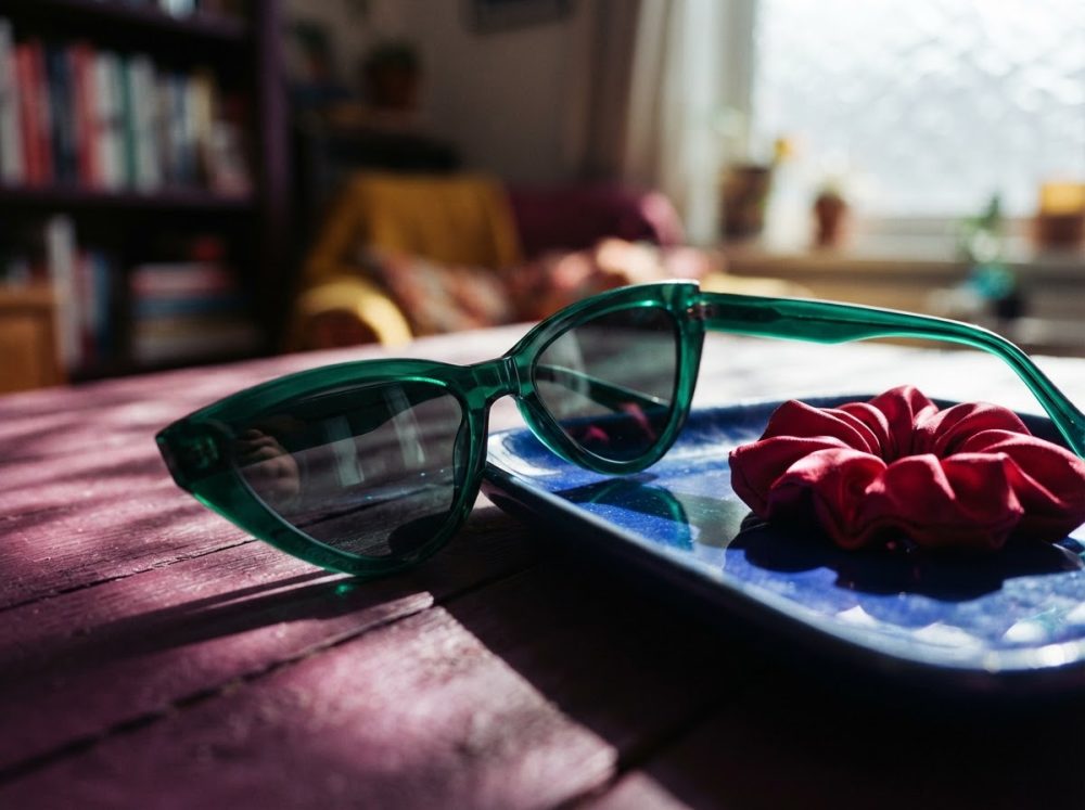 Óculos de sol verdes com scrunchie vermelho sobre bandeja azul numa superfície de madeira roxa e ambiente acolhedor