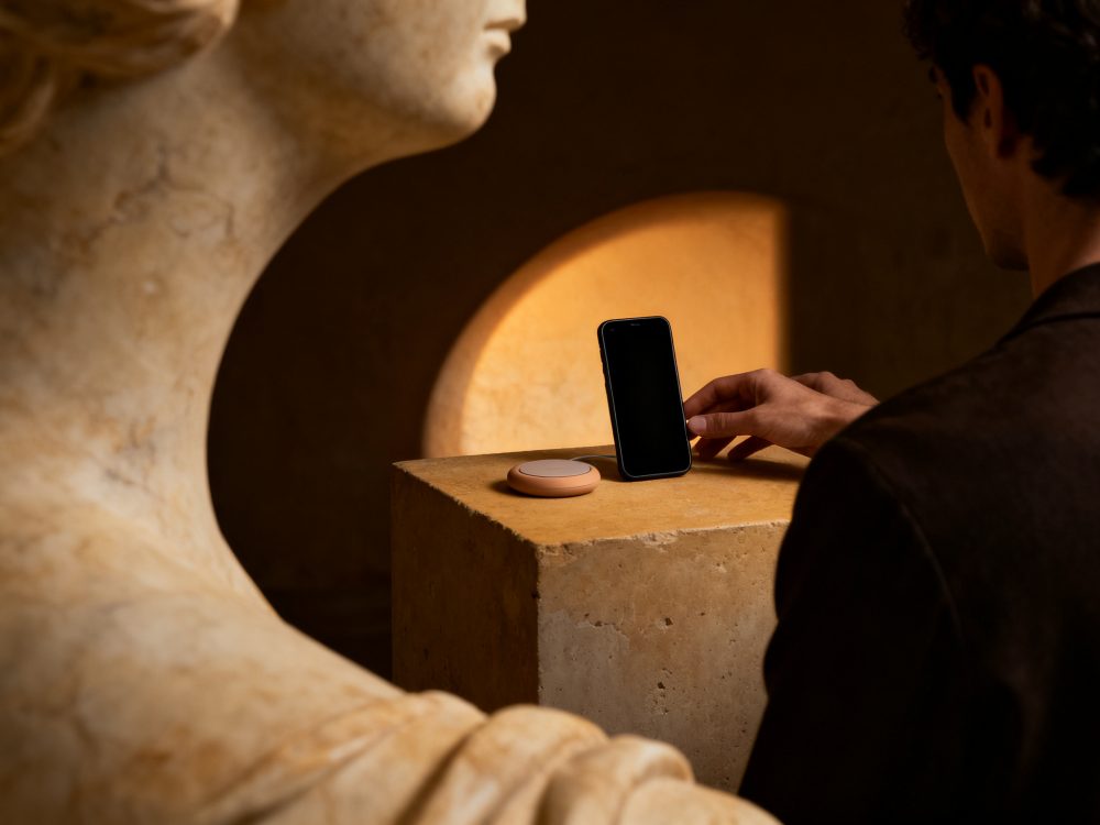Suporte para telemóvel preto com carregador wireless sobre pedestal de pedra com escultura clássica ao lado