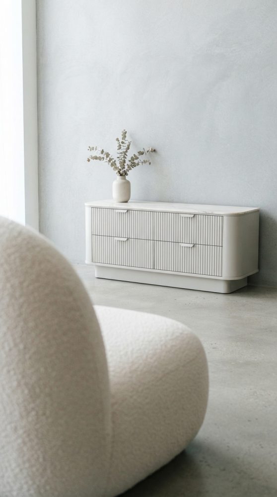 Cômoda branca minimalista com quatro gavetas e vaso com ramos secos, vista com cadeira creme em primeiro plano