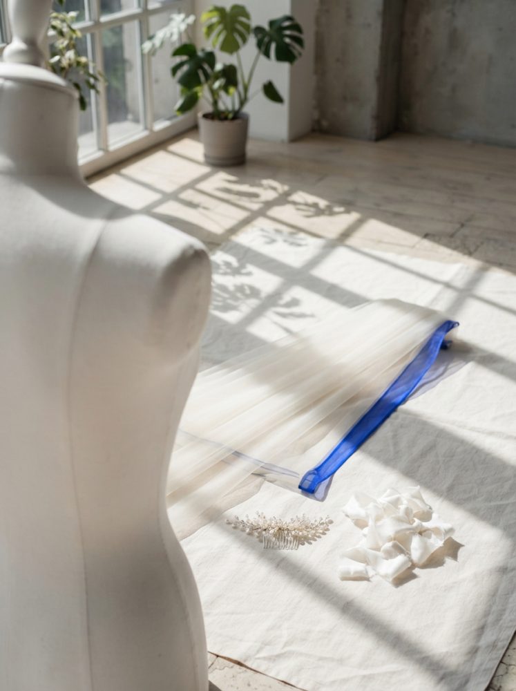 Véu de noiva branco com borda azul e florinhas ao lado de manequim num espaço iluminado por luz natural