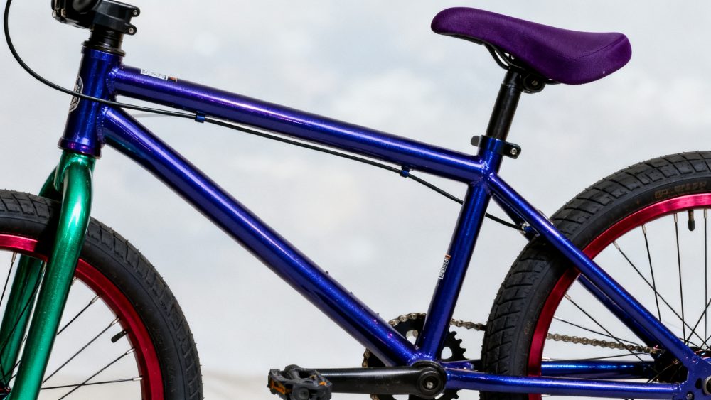 Bicicleta com estrutura azul metálica, assento roxo e rodas vermelhas sobre fundo neutro