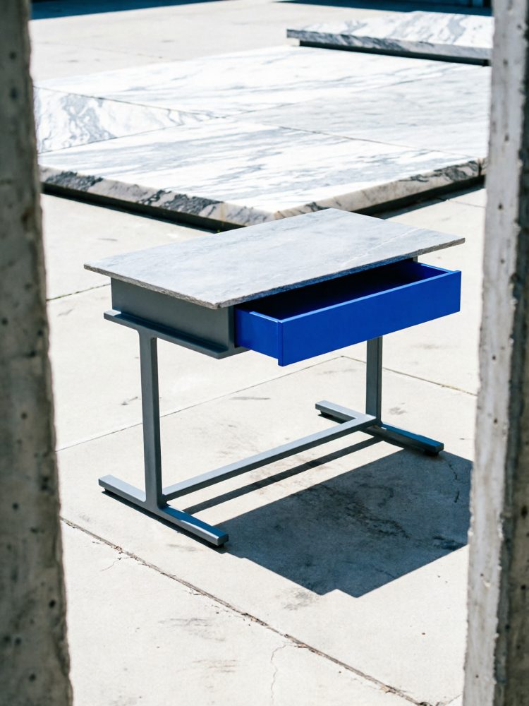 Mesa com tampo de pedra e gaveta azul sobre piso de cimento ao ar livre