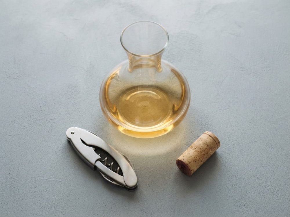 Decantador com vinho branco, saca-rolhas metálico e rolha sobre superfície cinza