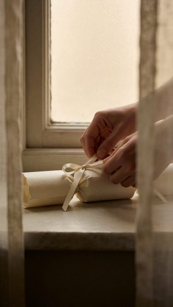 Mãos fechando cracker de Natal em papel creme com fita de cetim junto a janela
