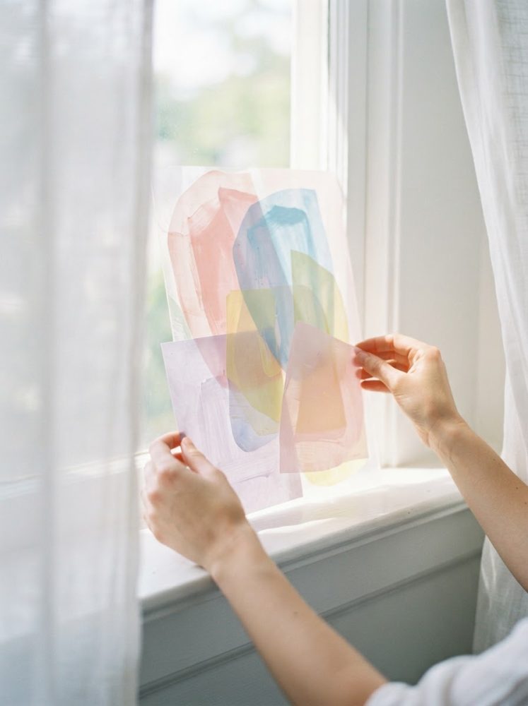 Mãos seguram composição de arte abstrata com papéis coloridos translúcidos junto a uma janela com luz suave
