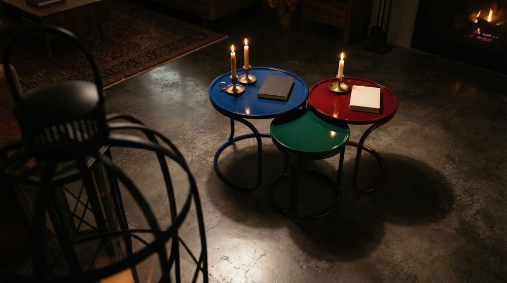 Conjunto de três mesas auxiliares redondas coloridas com velas acesas e livros numa sala com chão de cimento e lareira
