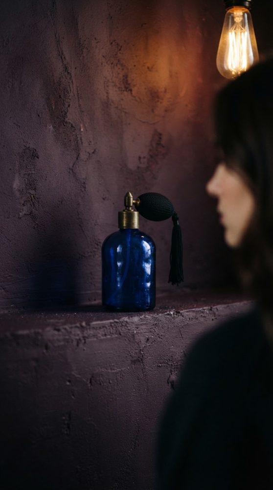 Frasco azul vintage de perfume com borrifador e luz quente ao fundo, com rosto desfocado feminine