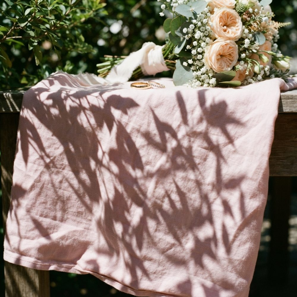 Bouquet de casamento com rosas e alianças sobre tecido rosa com sombras de folhas