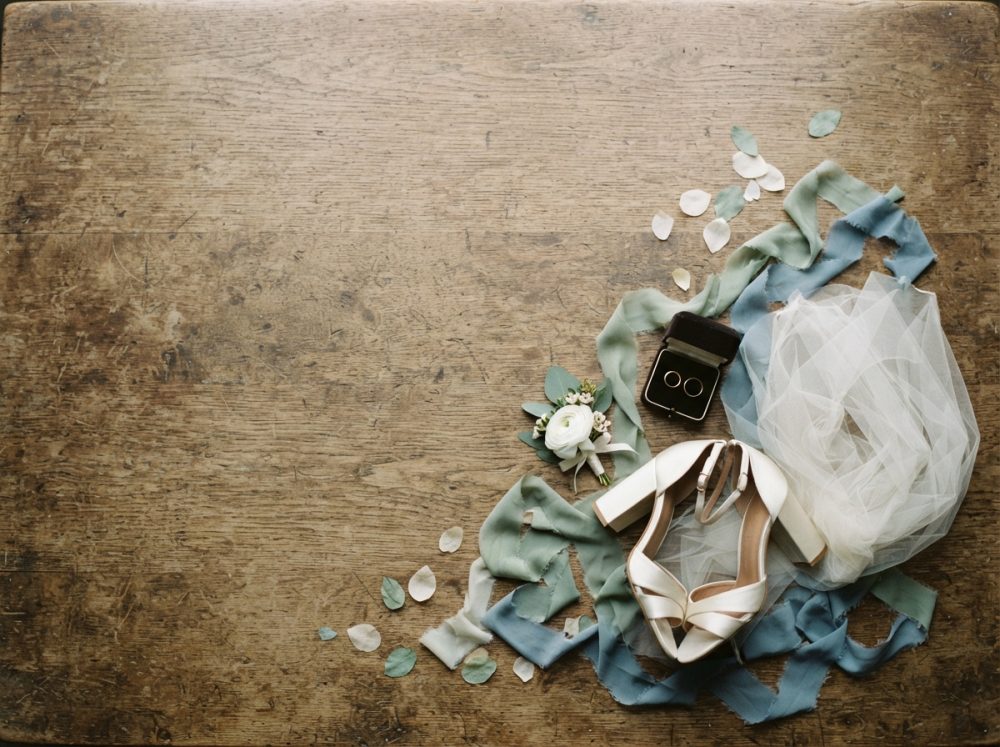 Sapatos de salto, alianças, flores e fitas sobre madeira rústica numa composição de casamento